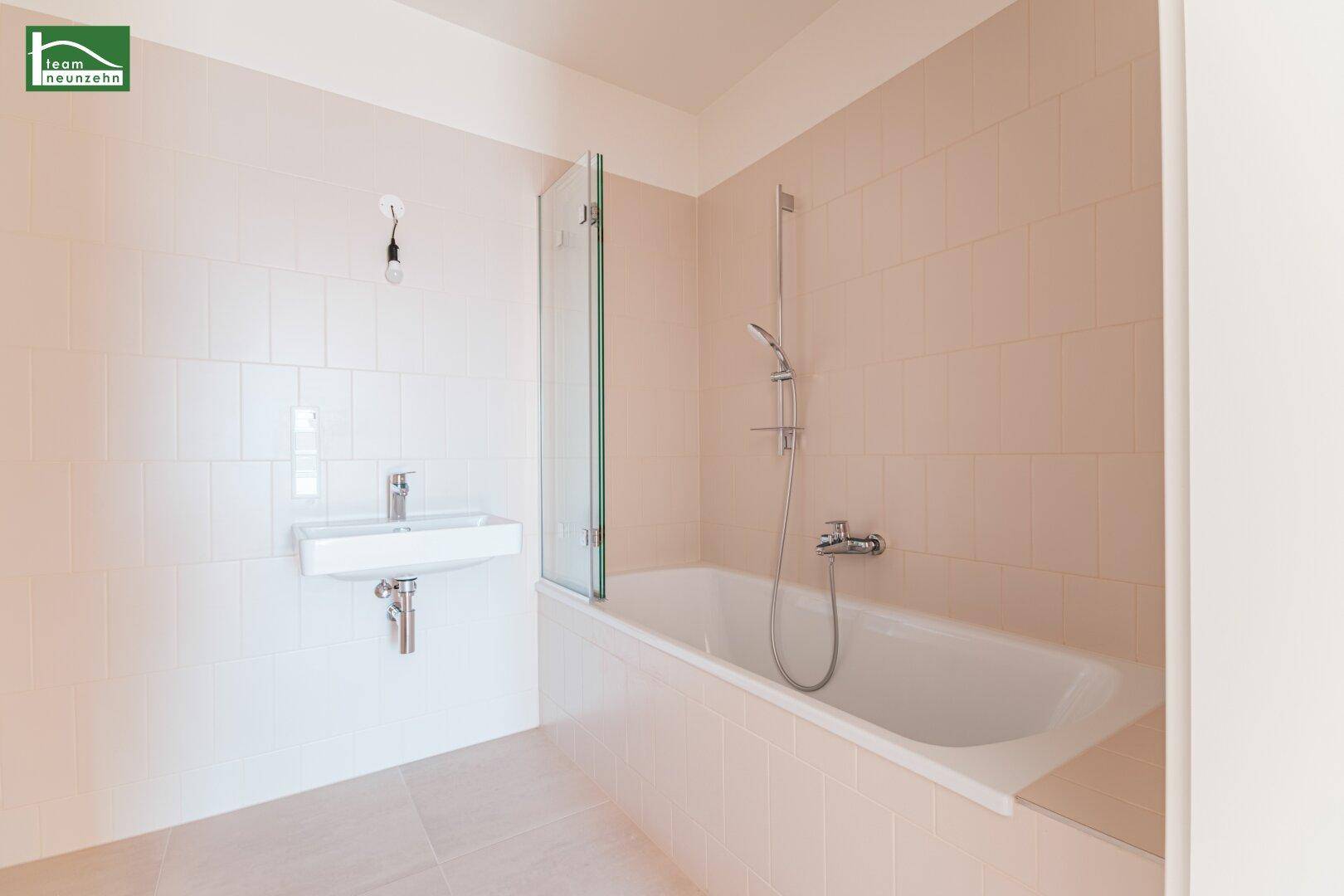 Badezimmer mit hellbeigen Fliesen, Badewanne mit Duschwand und modernem Waschbecken.