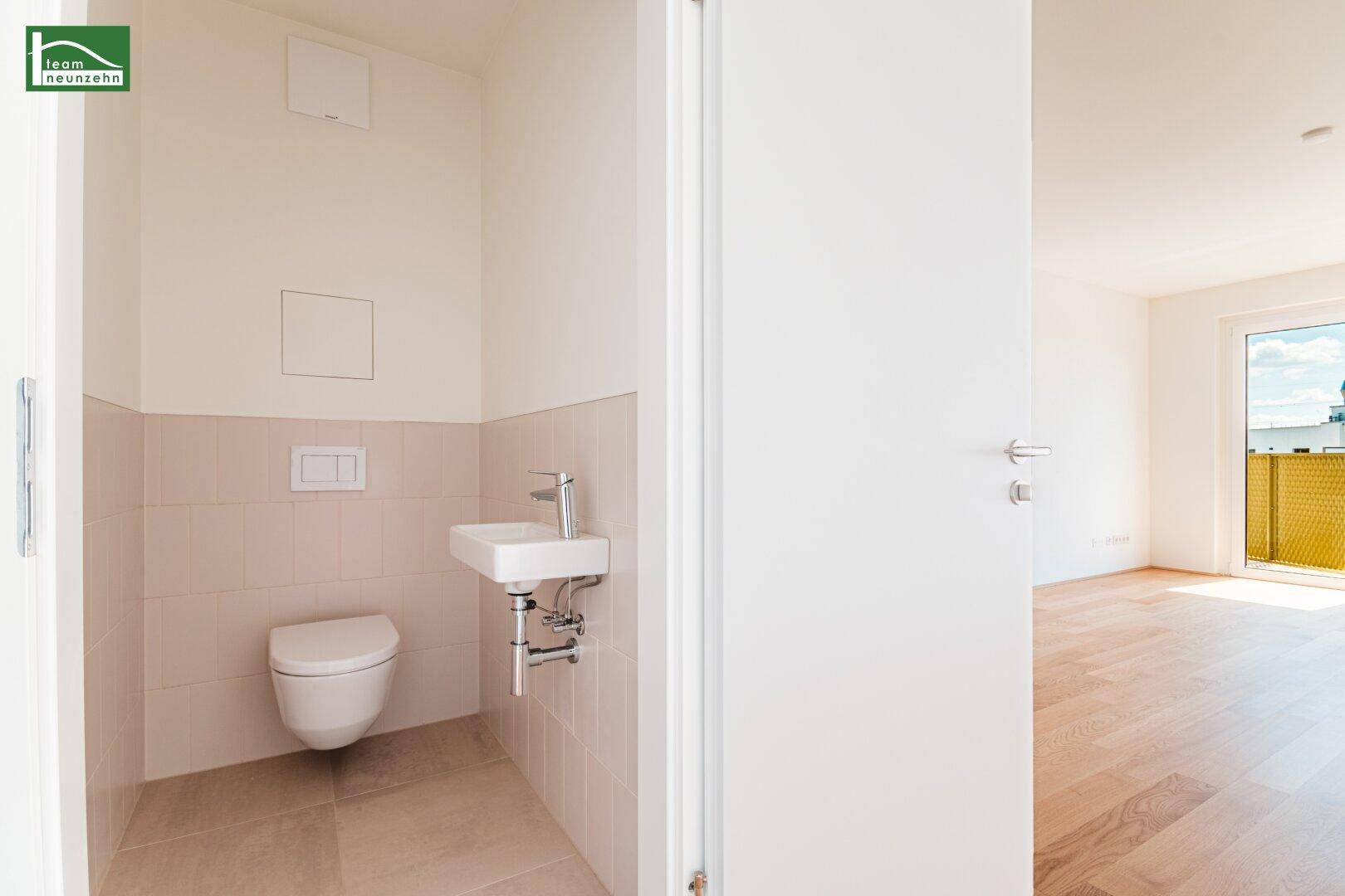 Separates WC mit wandhängender Toilette und kleinem Handwaschbecken, hell gefliest.