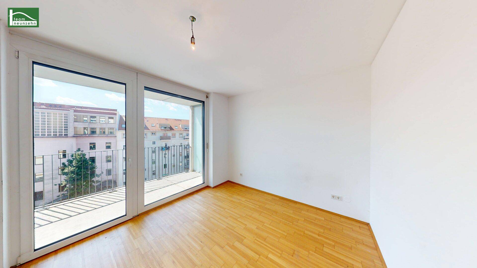 Helles Zimmer mit Holzboden und Zugang zu einem Balkon, bietet einen Blick auf umliegende Gebäude.