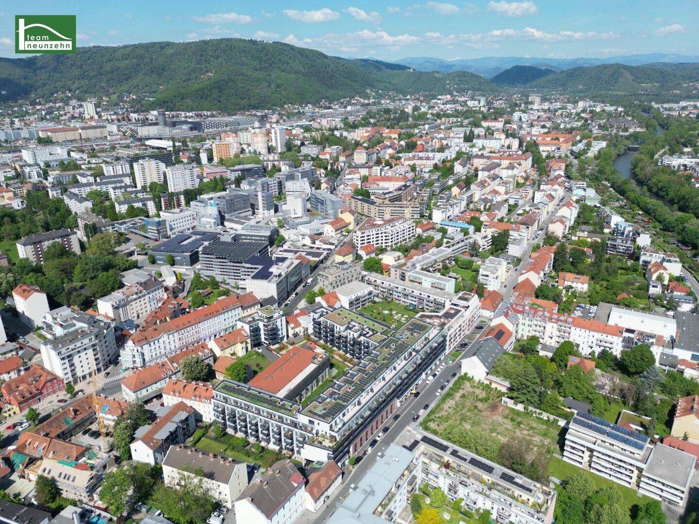 Luftaufnahme einer Stadt mit Wohngebäuden, Grünflächen und Bergen im Hintergrund an einem sonnigen Tag.