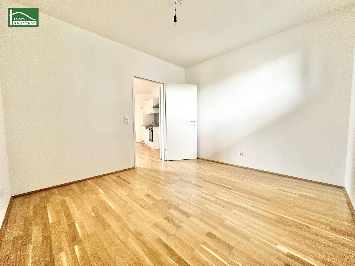 Geräumiges Zimmer mit Parkettboden und offener Tür zu einer dahinterliegenden Küche.