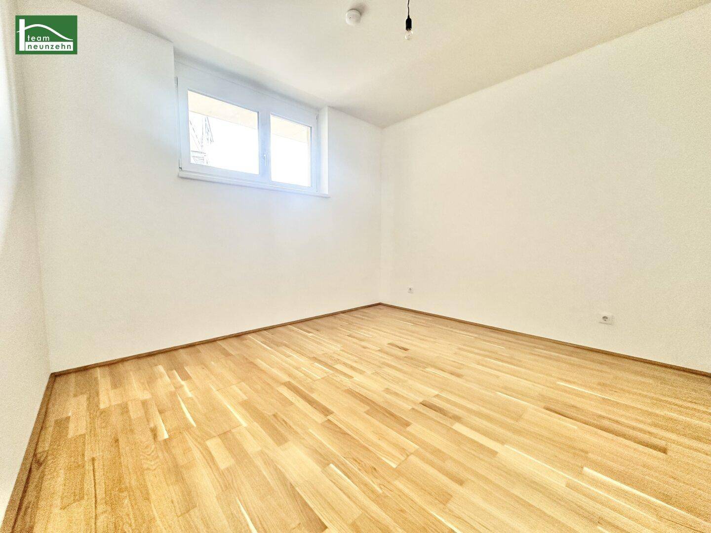 Helles, leeres Zimmer mit hellem Parkettboden und einem Fenster, das viel Tageslicht hereinlässt.