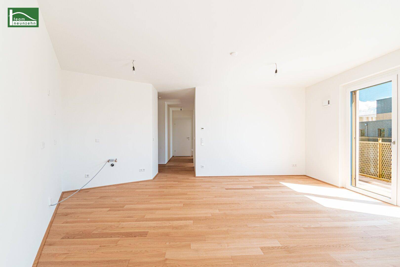 Heller, leerer Raum mit Holzboden und Zugang zum Balkon, führt in einen Flur.