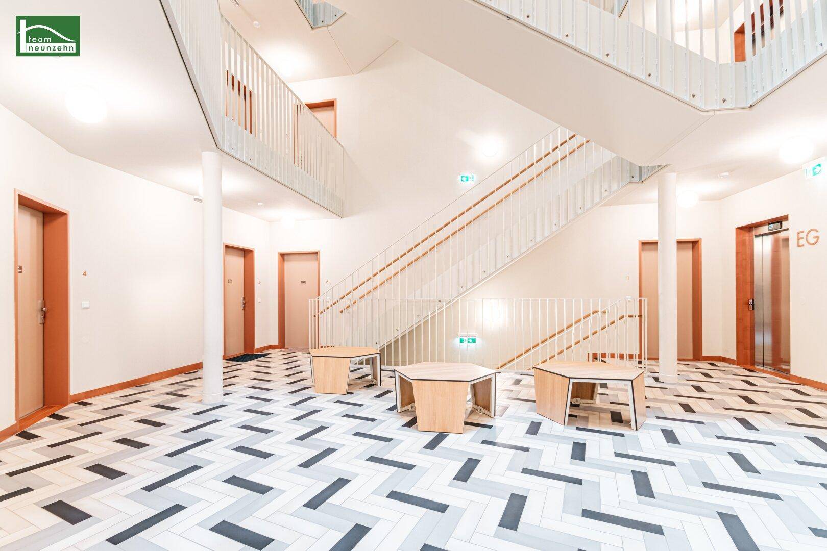 Heller Eingangsbereich mit modernem Treppenhaus, Aufzug und gemustertem Fliesenboden.