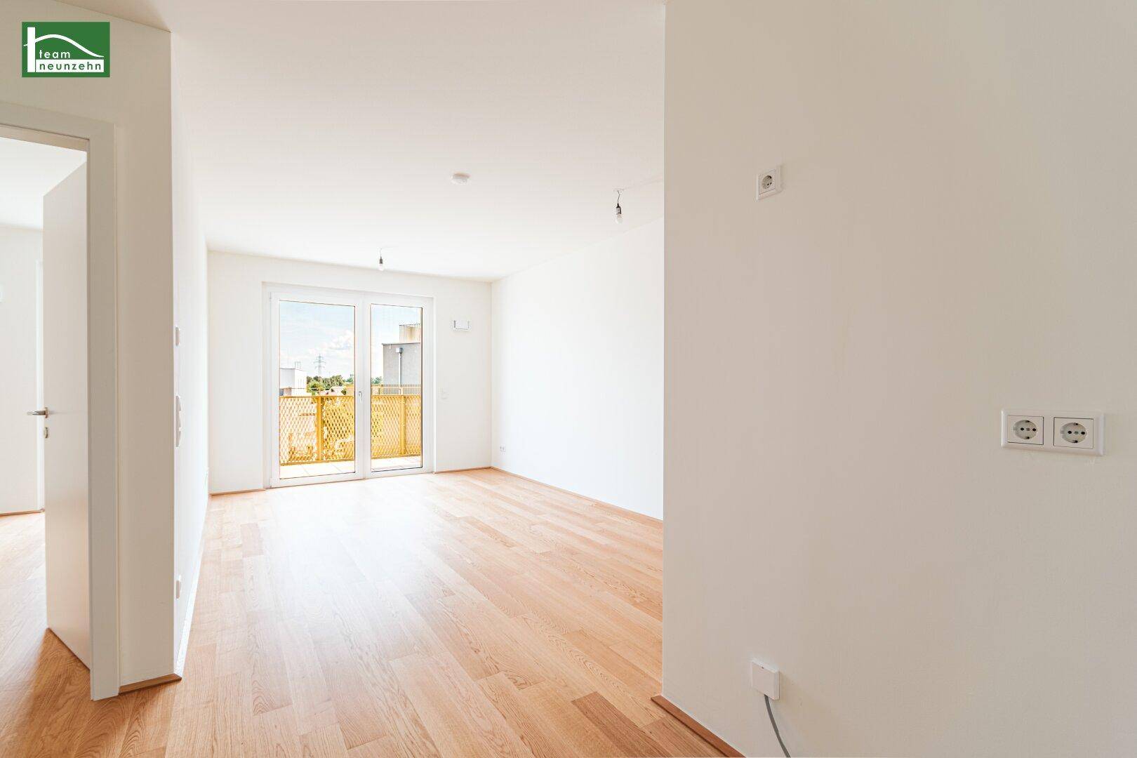 Heller, leerer Raum mit hellem Holzfußboden und Zugang zu einem sonnigen Balkon.