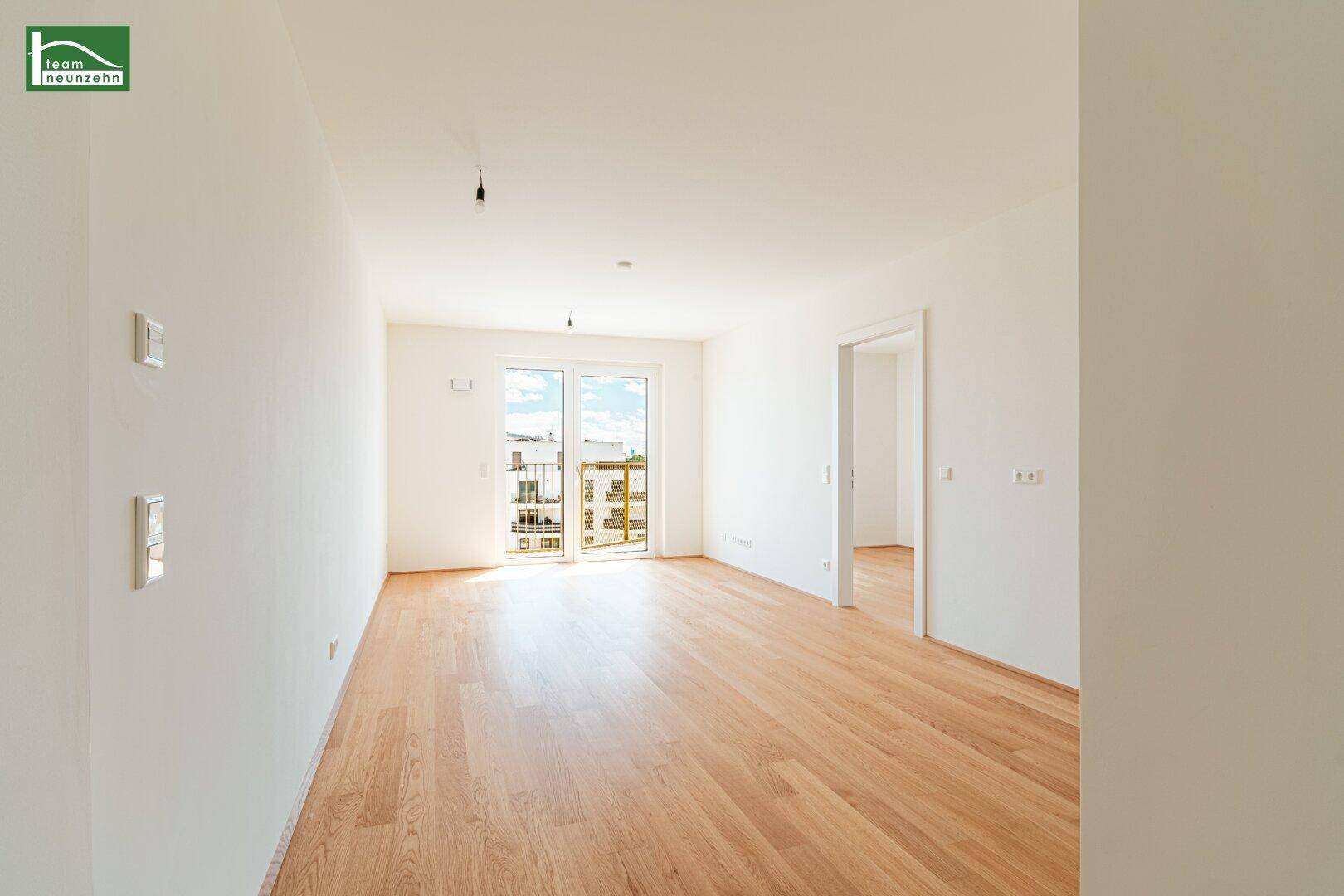 Lichtdurchfluteter Raum mit Holzboden und großen Fenstern, die Zugang zu einem Balkon bieten.