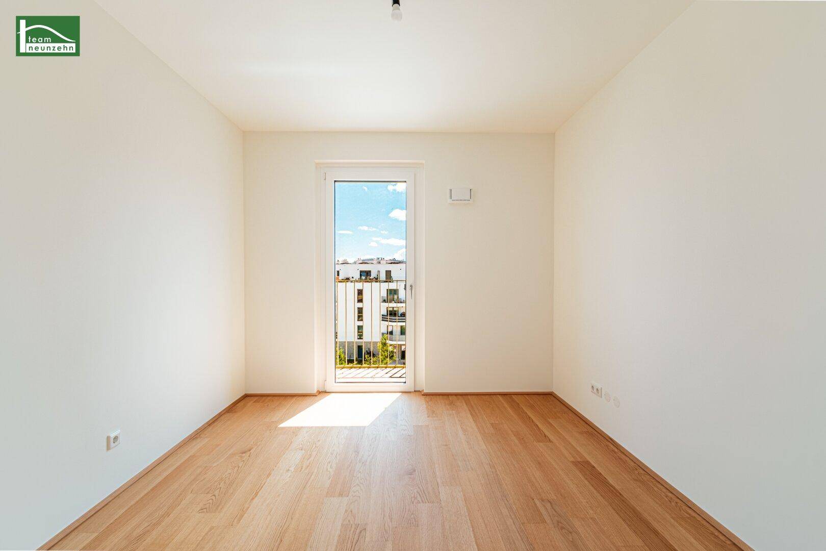 Helles, leeres Zimmer mit Holzboden und großem Fenster mit Blick auf die Umgebung.