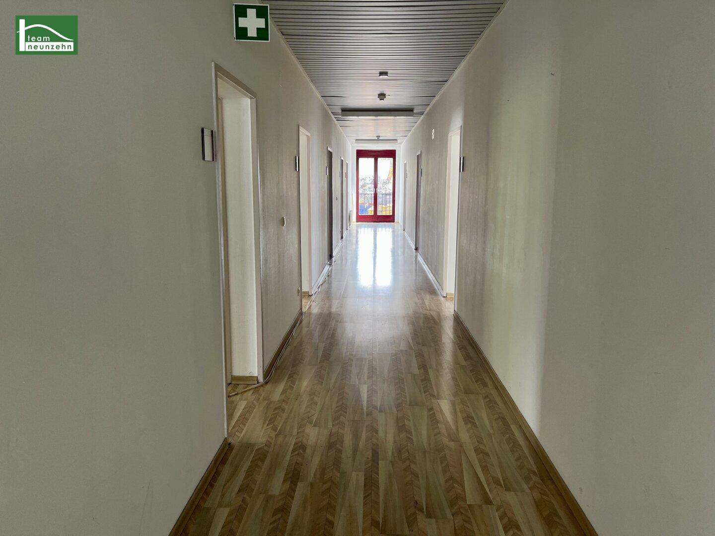 Langer, heller Flur mit Laminatboden, weißen Wänden und einem Notausgangsschild.