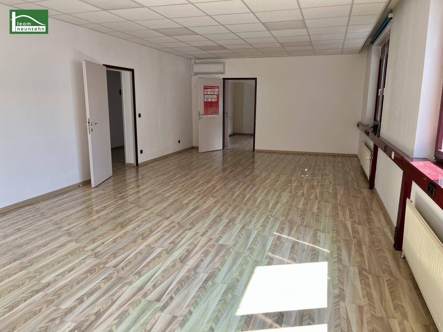 Großer Büroraum mit Laminatboden, Rasterdecke und Klimaanlage an der Wand.