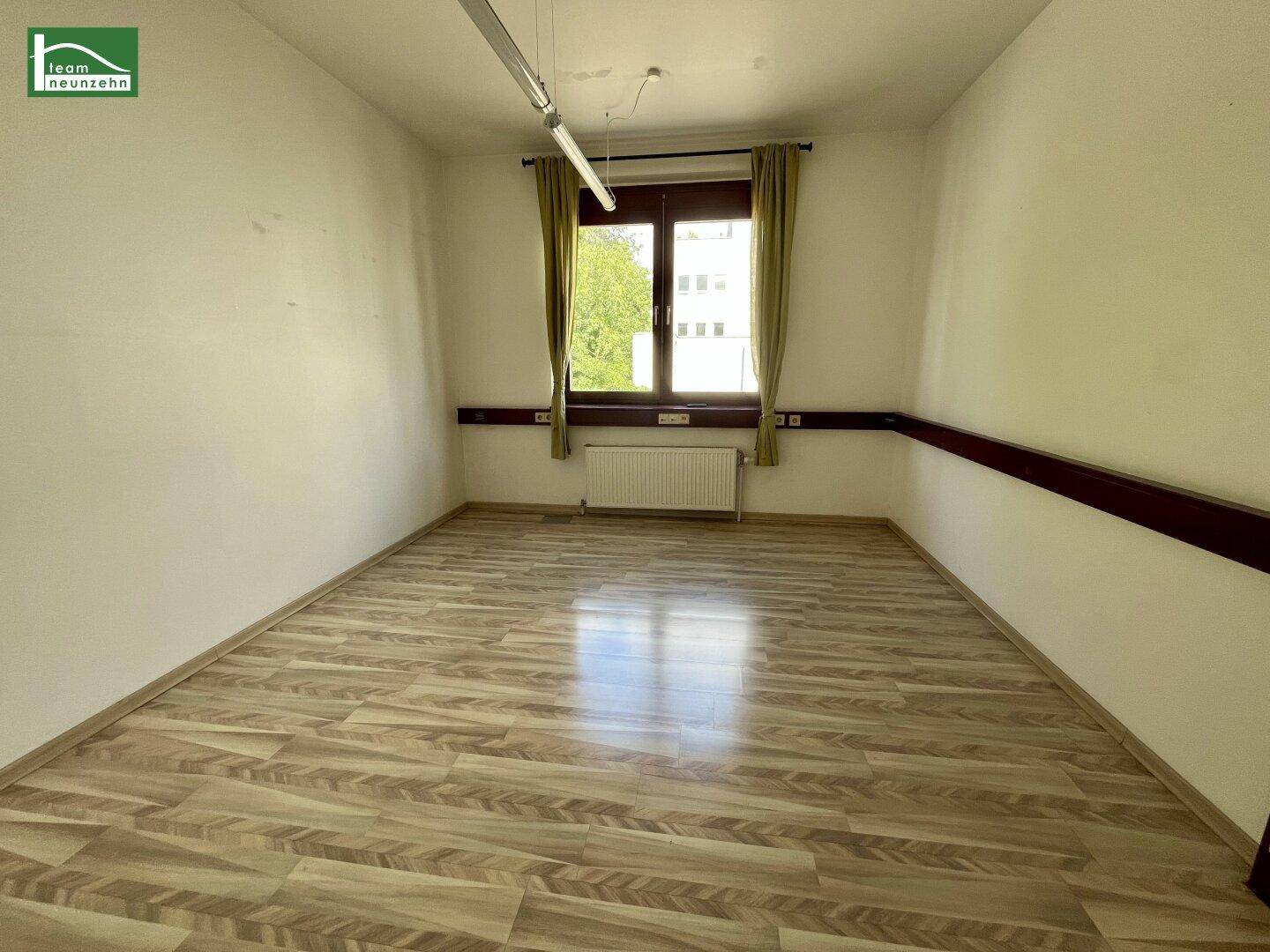 Heller Büroraum mit Laminatboden, einem Fenster und grünen Vorhängen.