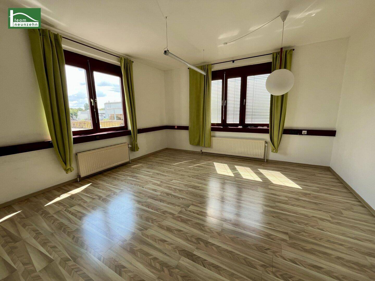 Heller Büroraum mit großen Fenstern, Holzoptik-Laminatboden und grünen Vorhängen.