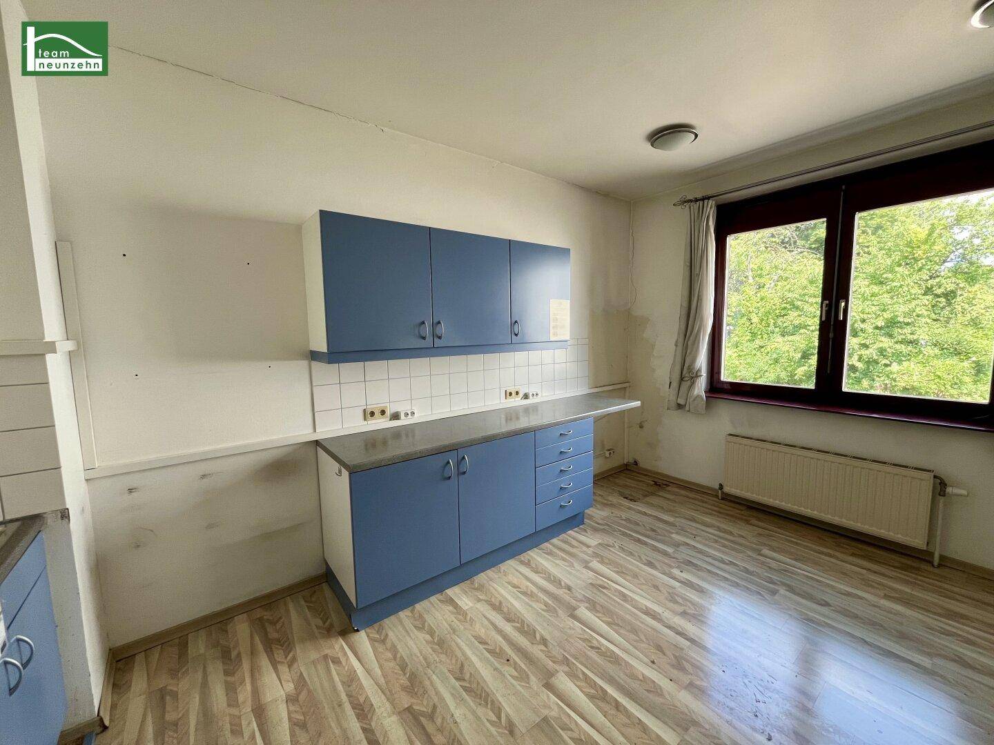Einfache Küche mit blauen Schränken, weißem Fliesenspiegel und Blick ins Grüne.
