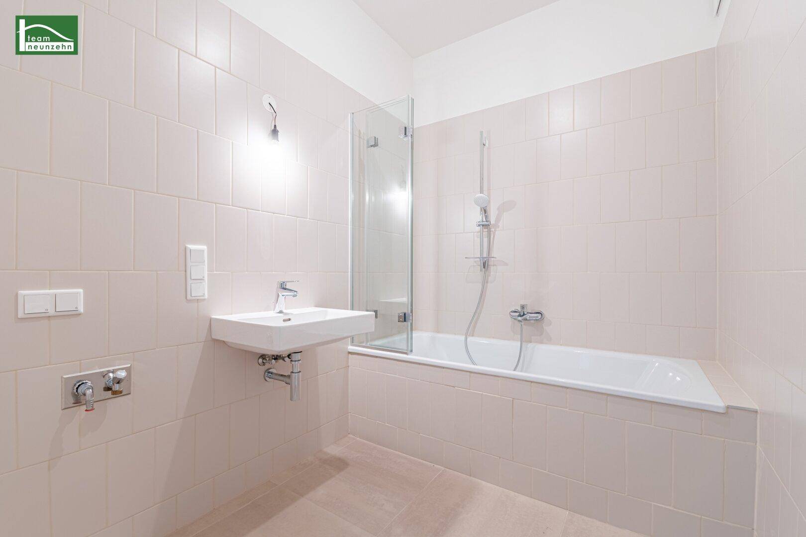 Modernes Badezimmer mit hellbeigen Fliesen, Badewanne mit Duschwand und wandhängendem Waschbecken.