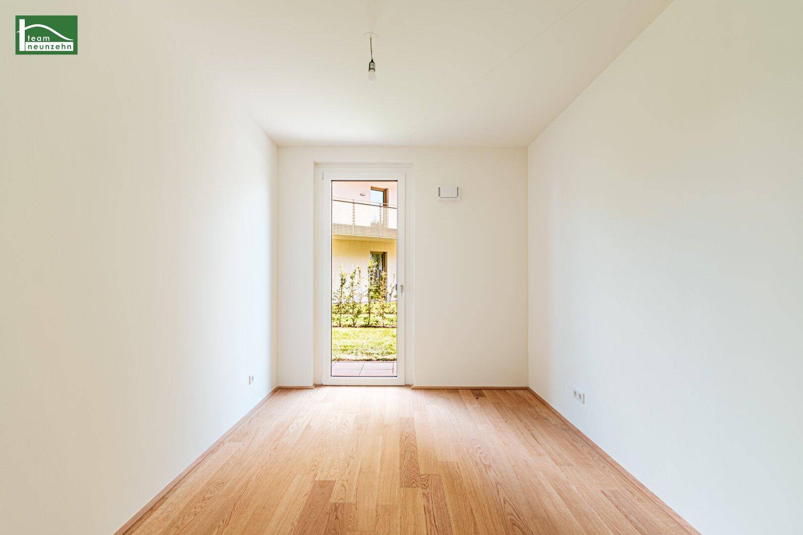 Leeres Zimmer mit hellem Parkettboden und Zugang zu einem kleinen Gartenbereich.