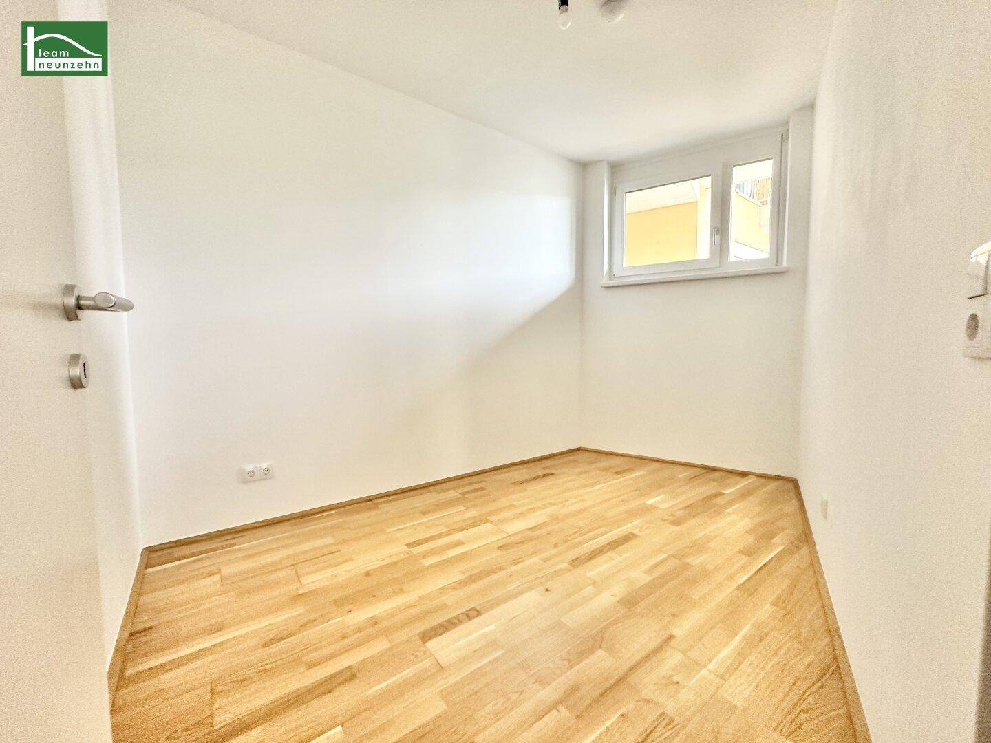Kleines, helles Zimmer mit Parkettboden, weißen Wänden und einem Fenster mit Rollladen.