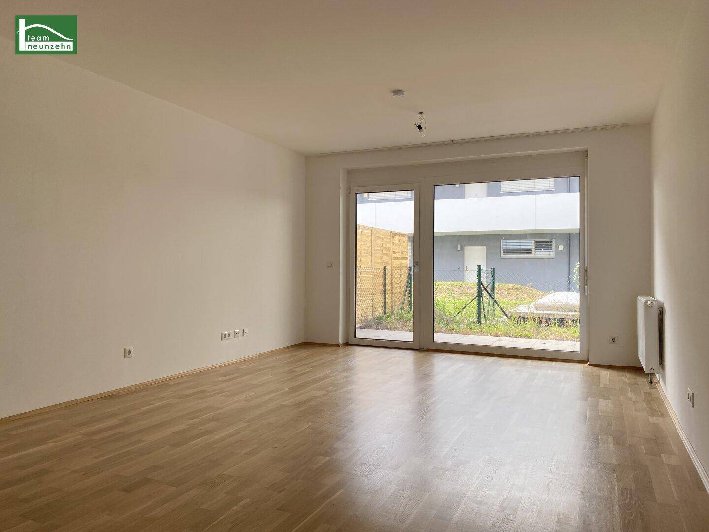 Helles, leeres Wohnzimmer mit Parkettboden und Zugang zu einem kleinen Außenbereich.