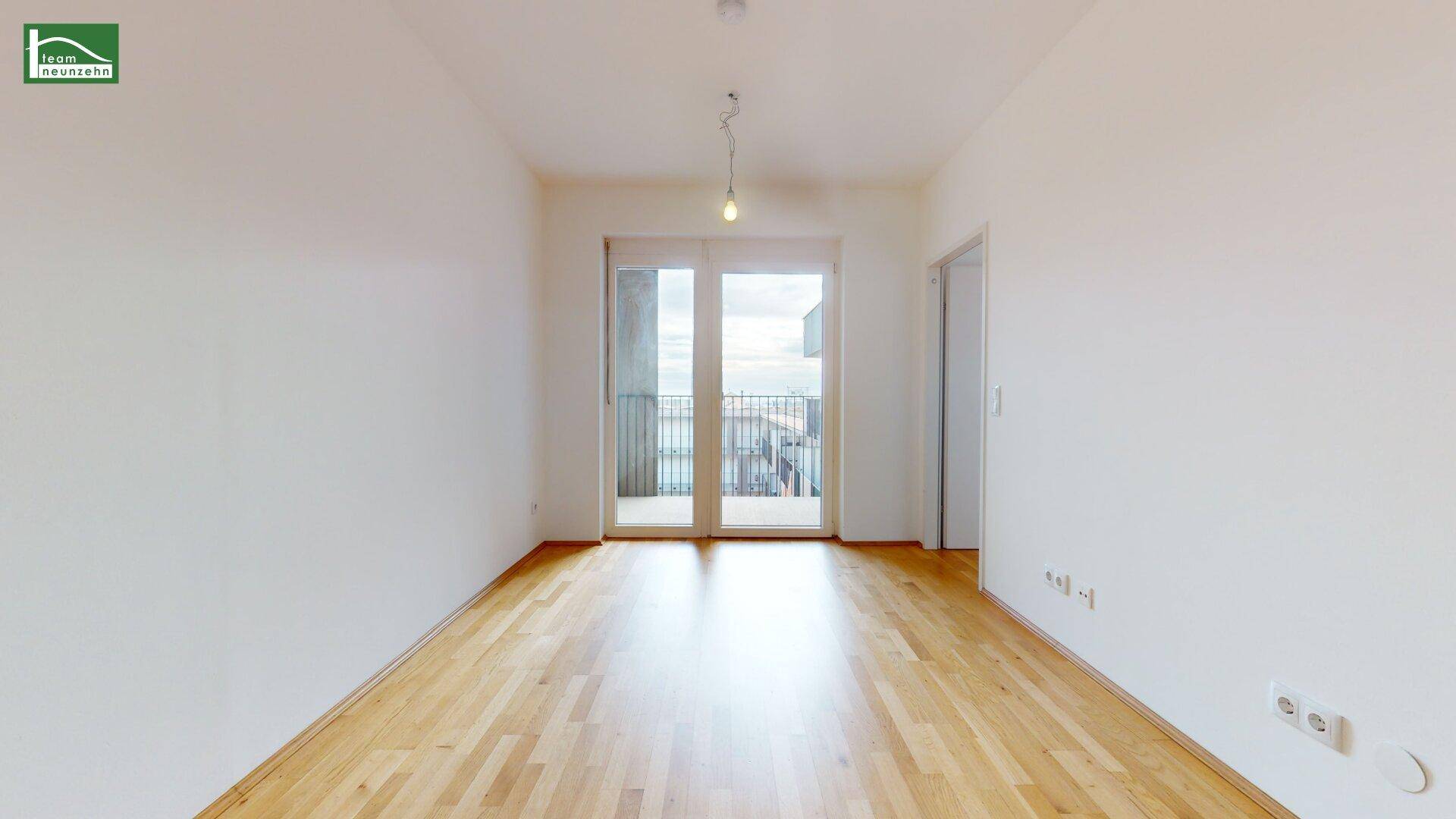 Geräumiger, unmöblierter Raum mit hellem Holzboden und großen Fenstern zum Balkon.