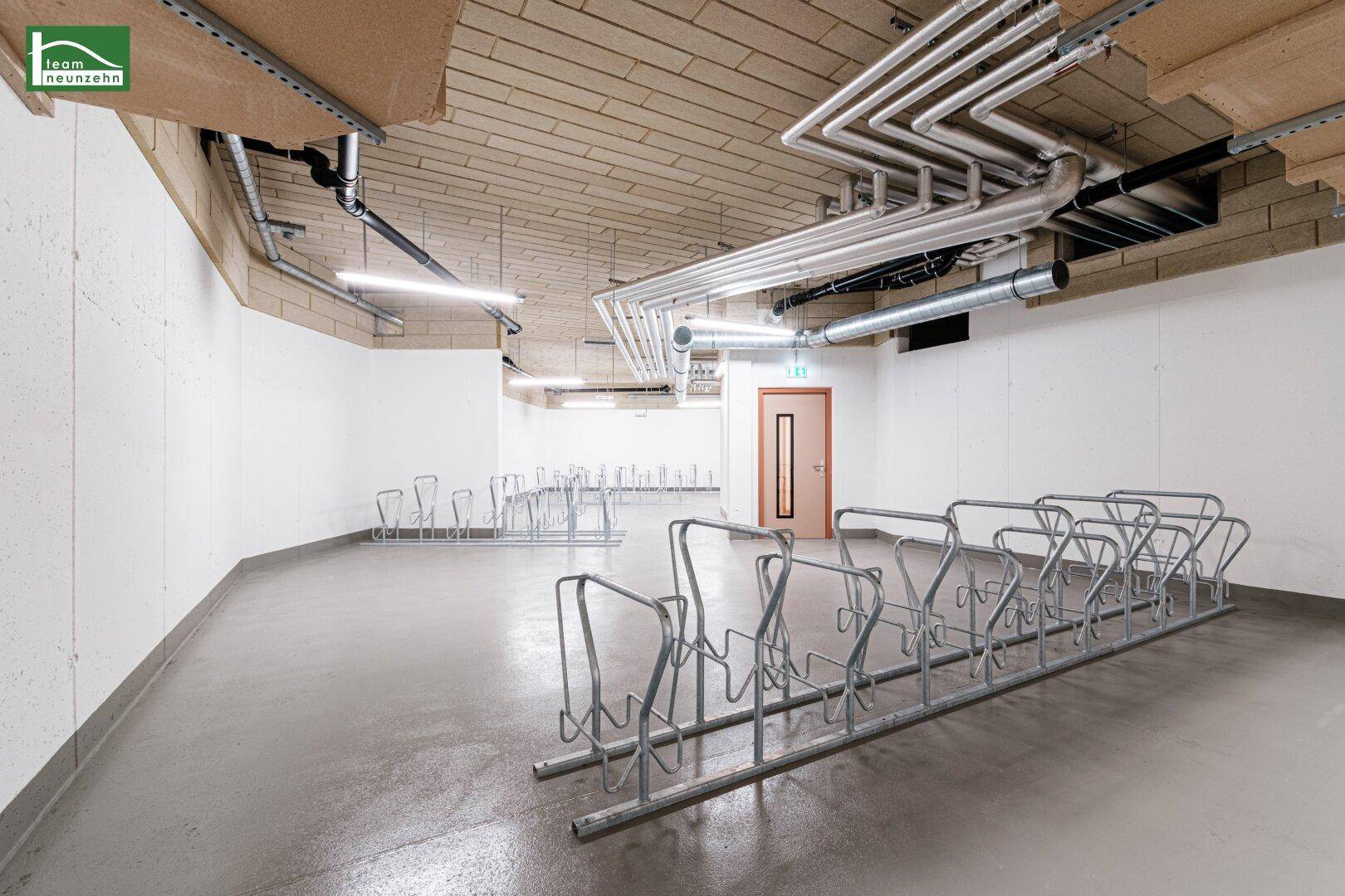 Großer Fahrradraum im Keller mit zahlreichen Fahrradständern und sichtbaren Rohrleitungen an der Decke.