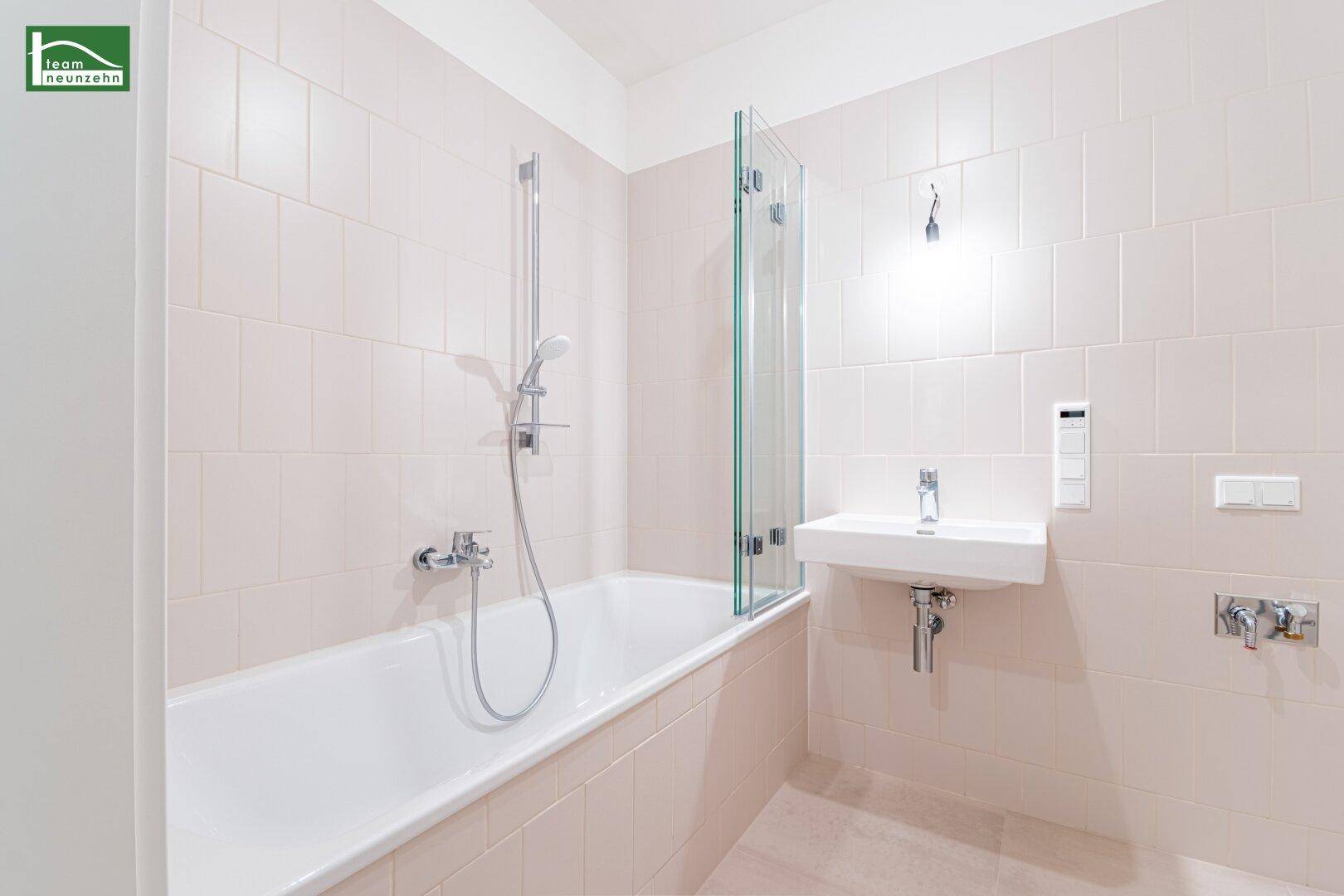 Modernes Badezimmer mit Badewanne, Duschwand und hellem Einzelwaschbecken, in beigen Fliesen gehalten.