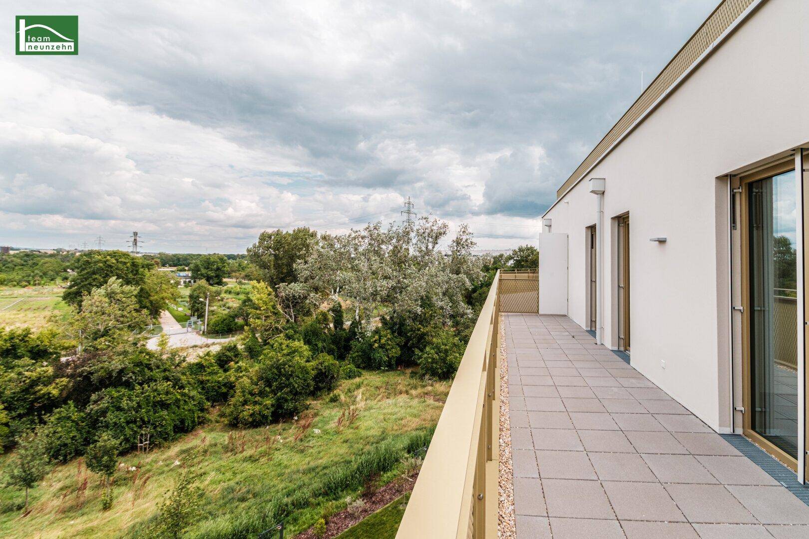 Langer Balkon mit Fliesenboden und Blick ins Grüne unter einem bewölkten Himmel.