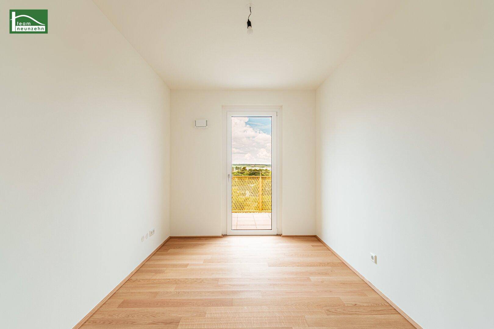 Helles Zimmer mit Holzboden und einer Tür, die zu einem Balkon oder einer Terrasse führt.