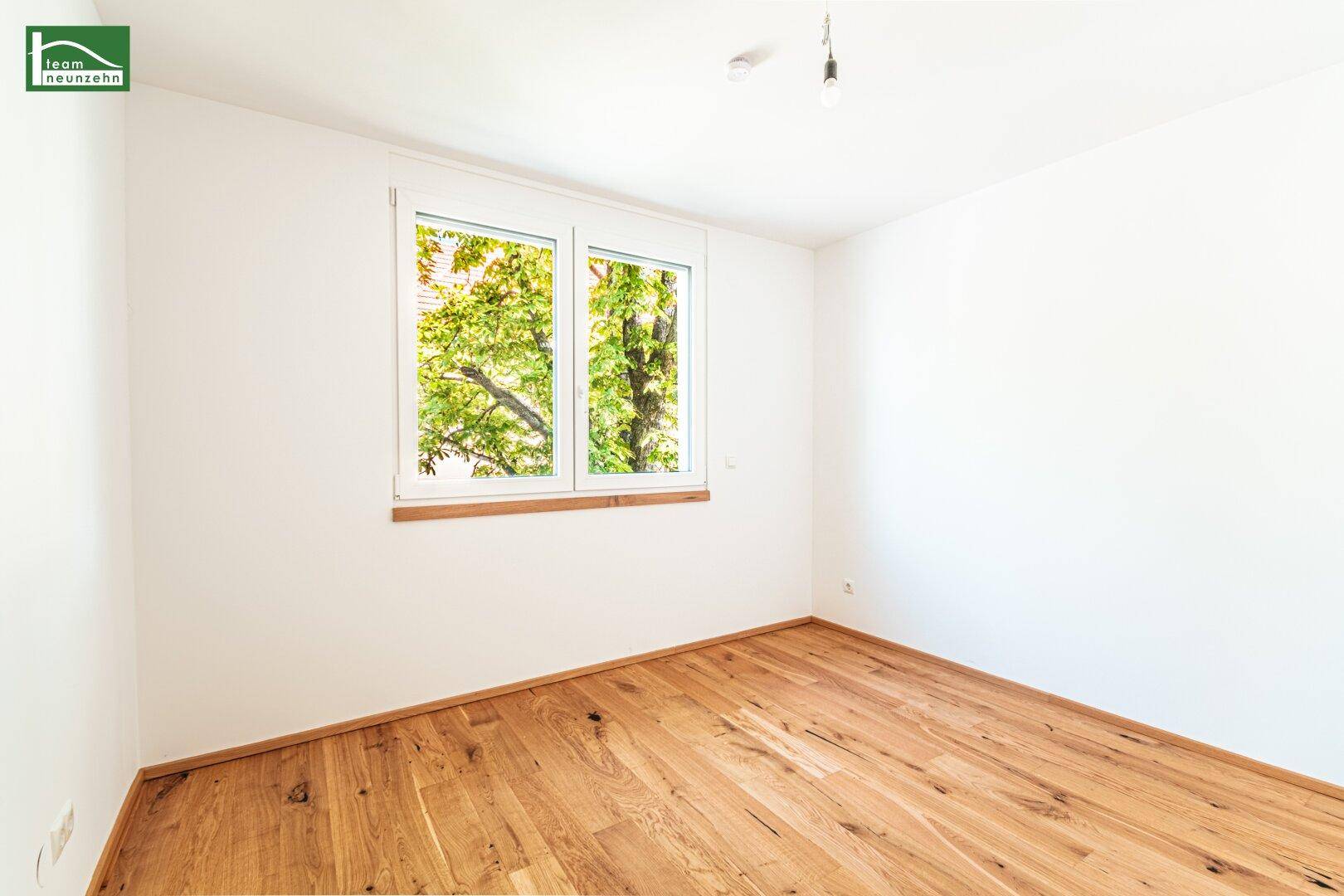 Helles, leeres Zimmer mit Holzboden und Fenster, das einen Blick ins Grüne bietet.