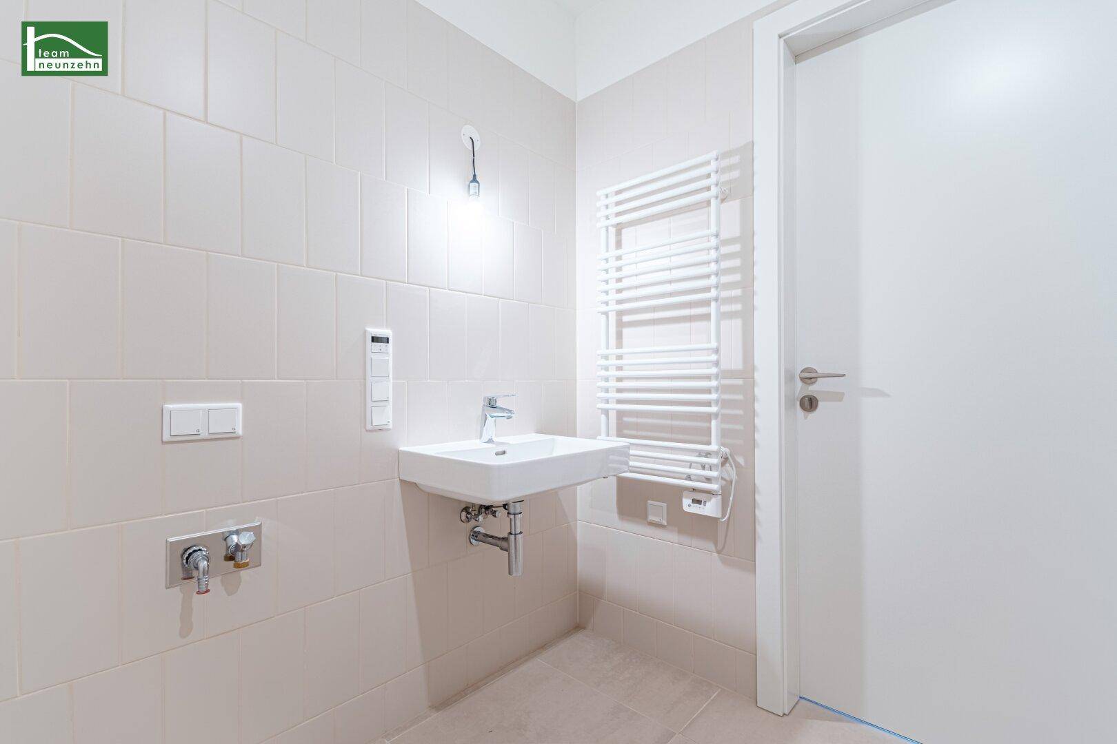 Helles Badezimmer mit wandmontiertem Waschbecken, beigen Fliesen und einem praktischen Handtuchheizkörper.