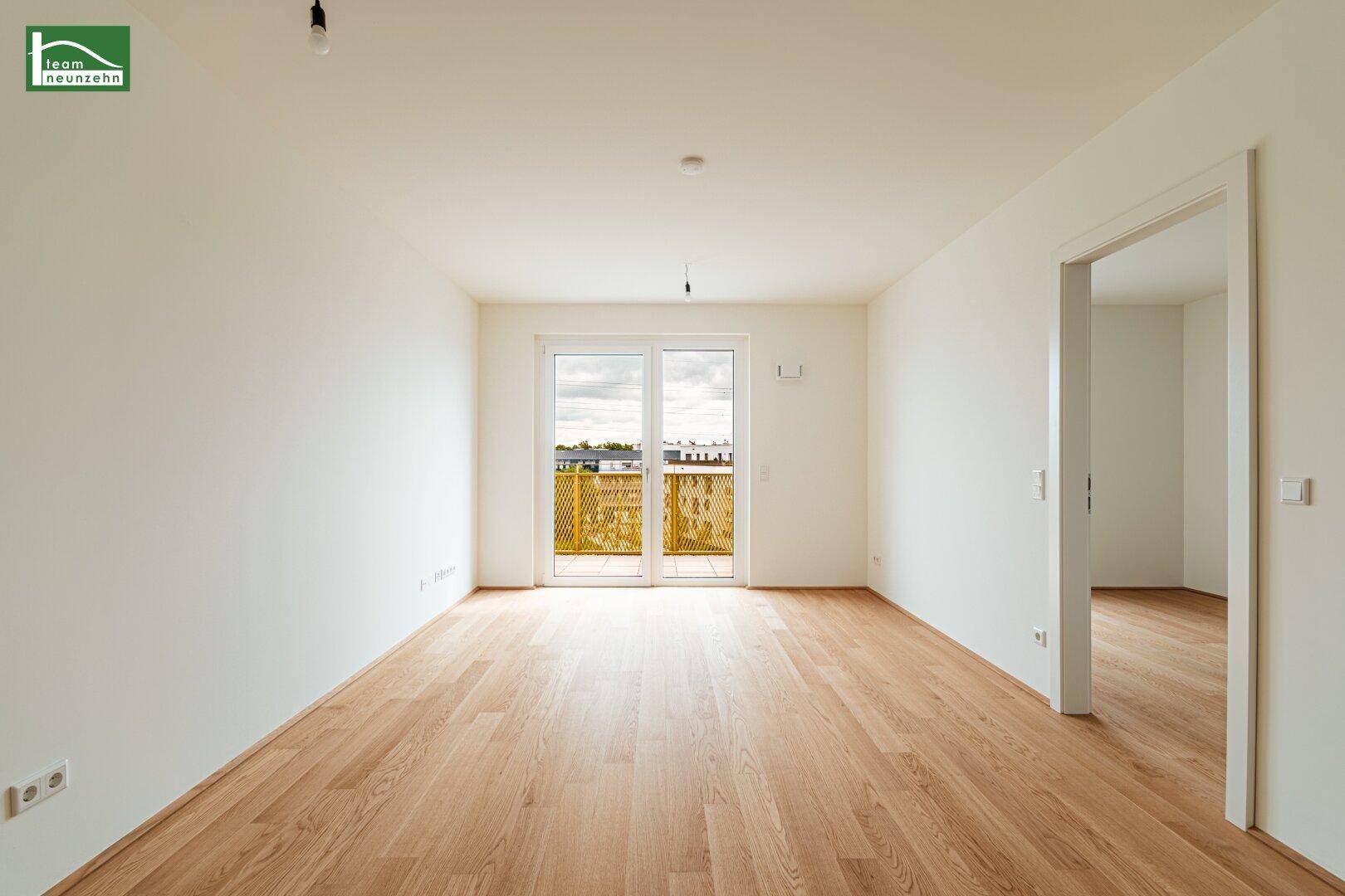 Helles, leeres Wohnzimmer mit Holzboden und Zugang zu einem Balkon.