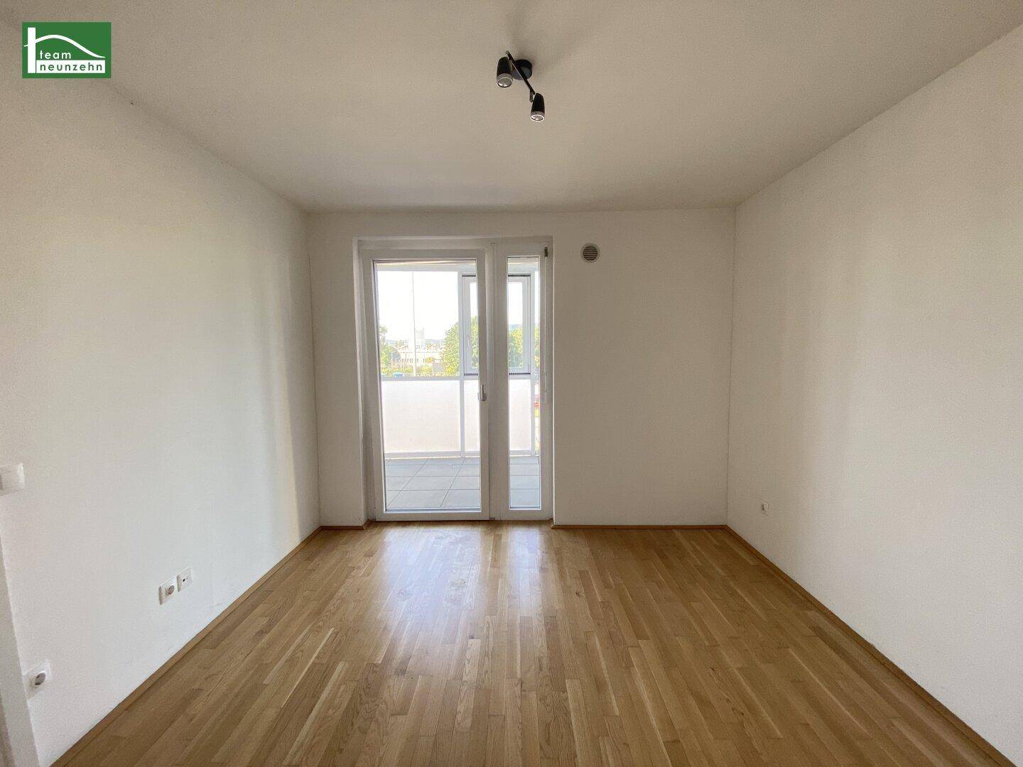 Leeres Zimmer mit hellem Parkettboden und großen Fenstern, die Zugang zu einem Balkon bieten.