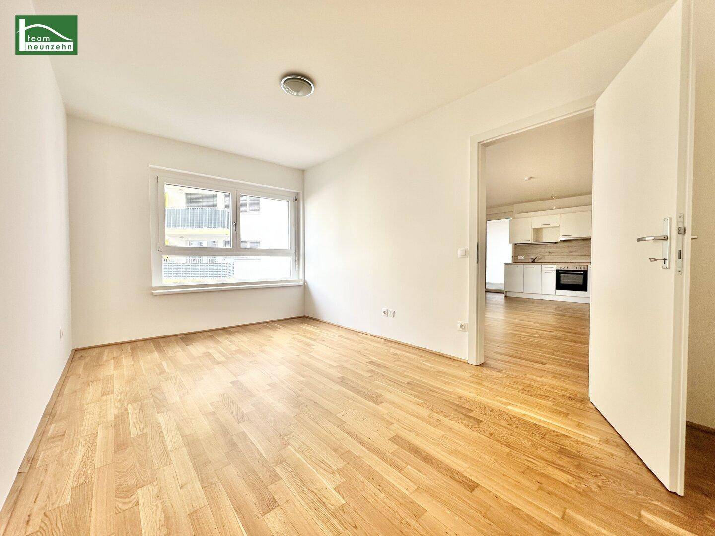 Helles, leeres Zimmer mit Parkettboden und Zugang zu einer modernen Küche.