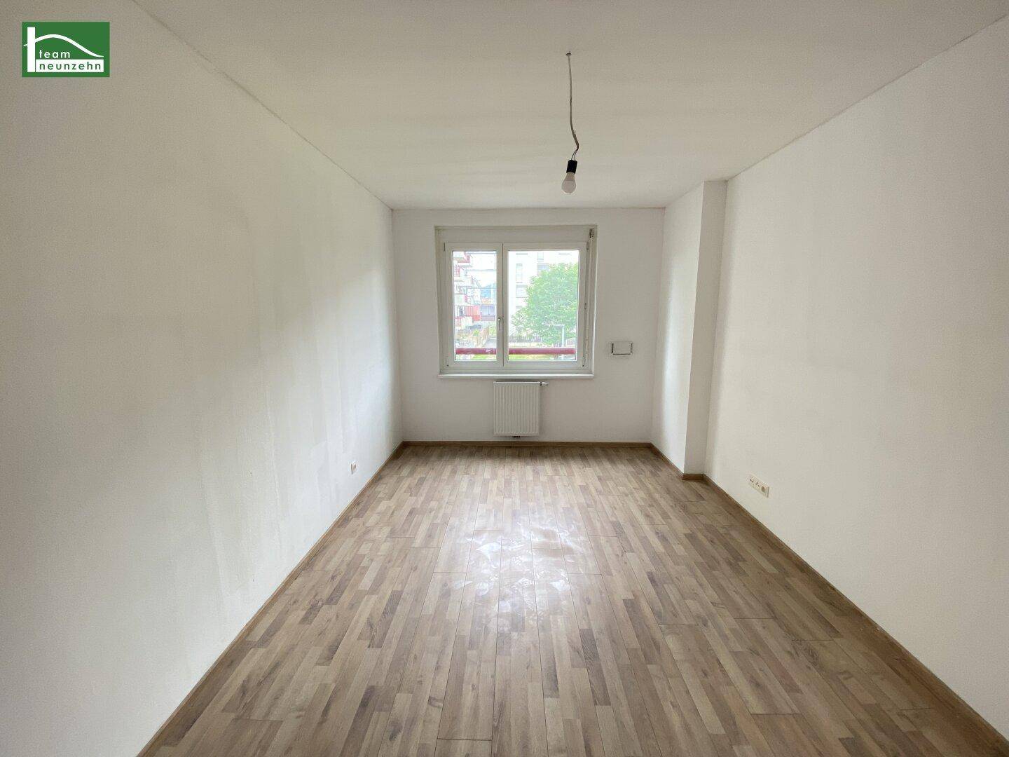 Helles, leeres Zimmer mit Parkettboden und einem Fenster, das Tageslicht hereinlässt.
