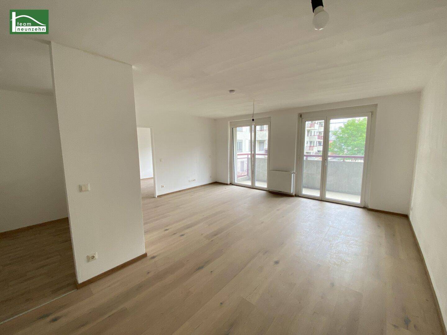 Geräumiger, unmöblierter Wohnbereich mit hellem Holzboden und Zugang zu einem Balkon.