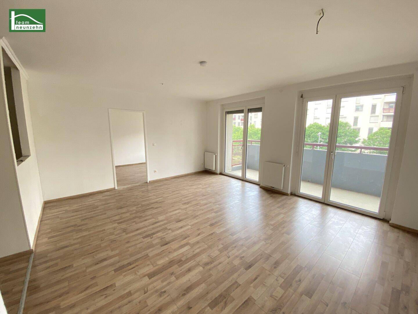 Helles, leeres Wohnzimmer mit Laminatboden und Zugang zu einem Balkon.