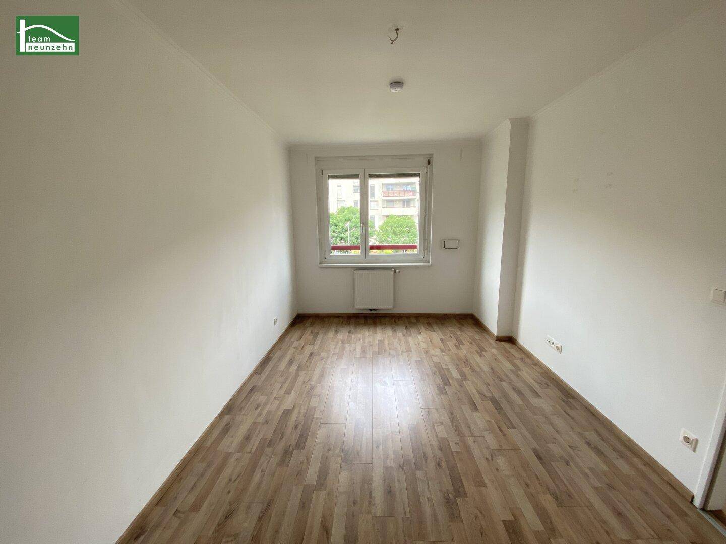 Leeres Zimmer mit Laminatboden, weiß gestrichenen Wänden und einem Fenster.