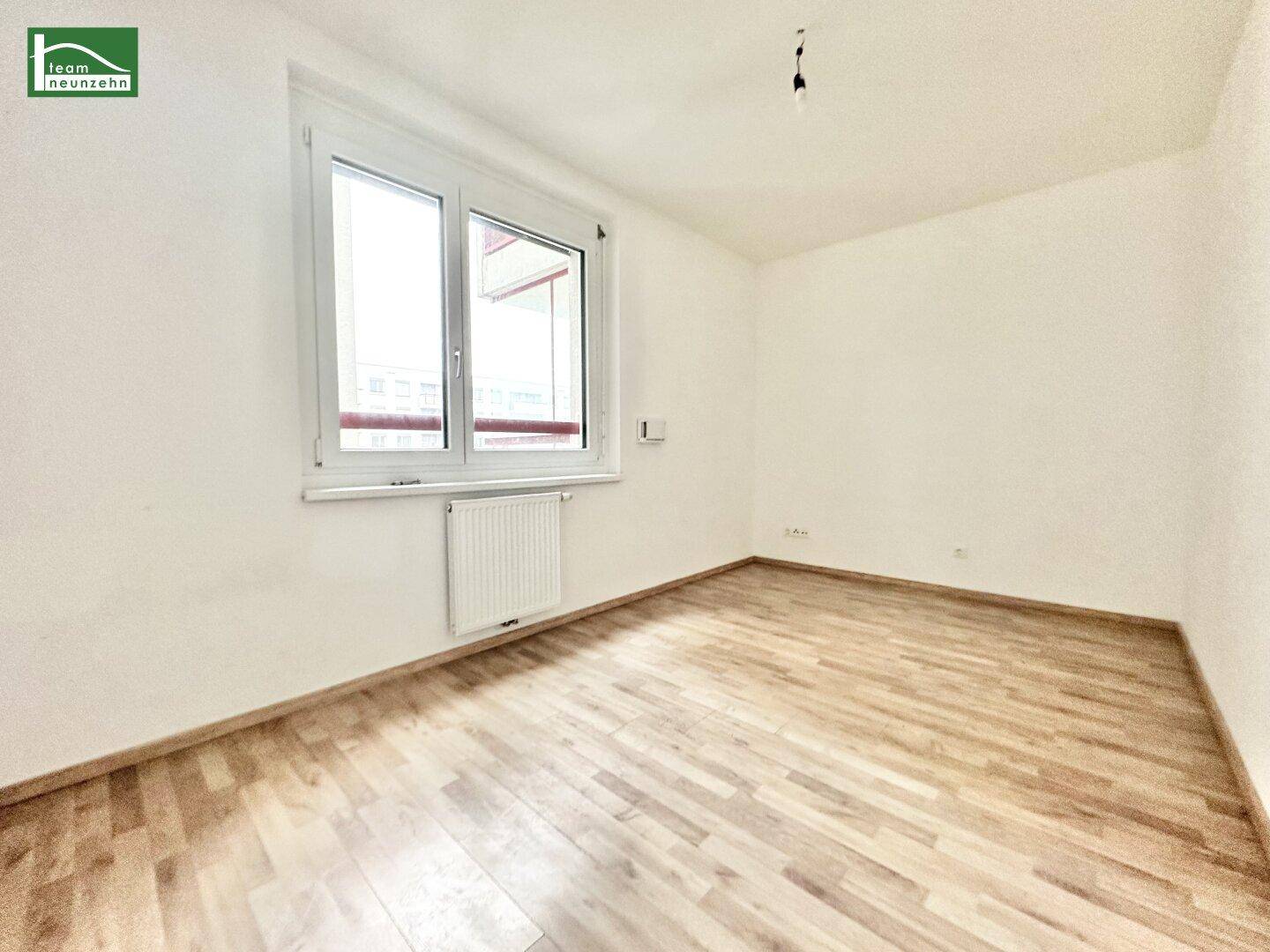 Leeres Zimmer mit Holzboden und großem Fenster, das viel natürliches Licht hereinlässt.