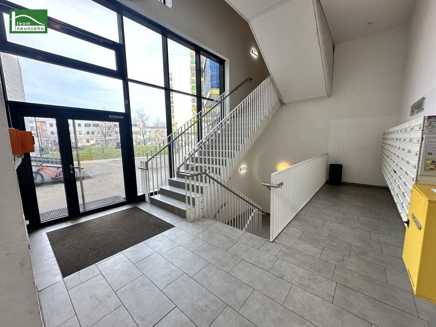 Eingangsbereich mit Treppe, großen Glastüren und Briefkästen in einem modernen Gebäude.