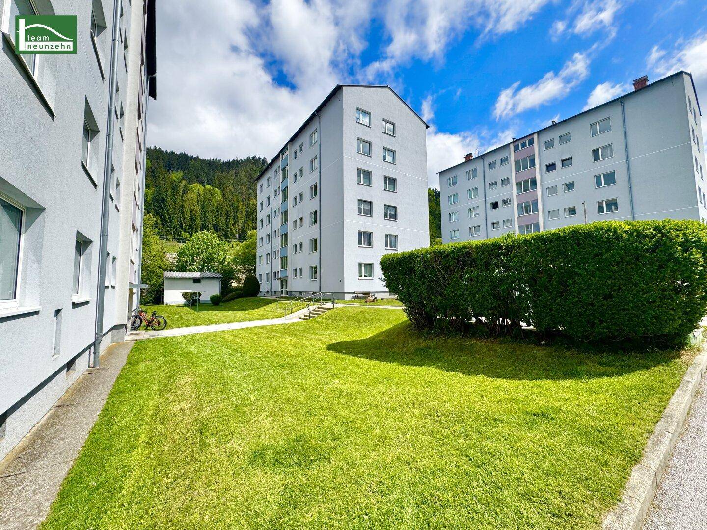Wohngebäude mit gepflegter Rasenfläche, Hecke und Blick auf die bewaldeten Berge.