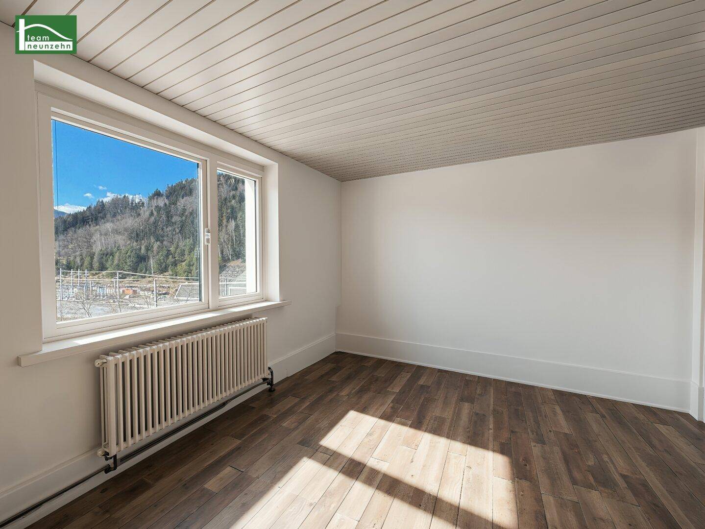 Helles Zimmer mit Holzboden, Holzdecke und weitem Blick auf die bewaldeten Hügel.