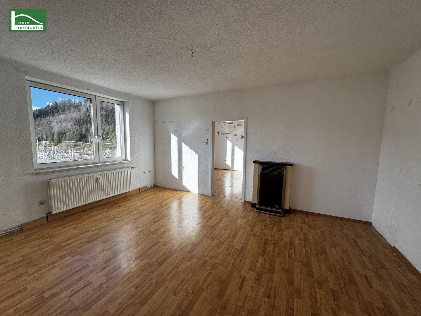 Geräumiges Wohnzimmer mit Parkettboden, großem Fenster und einem Ofen.