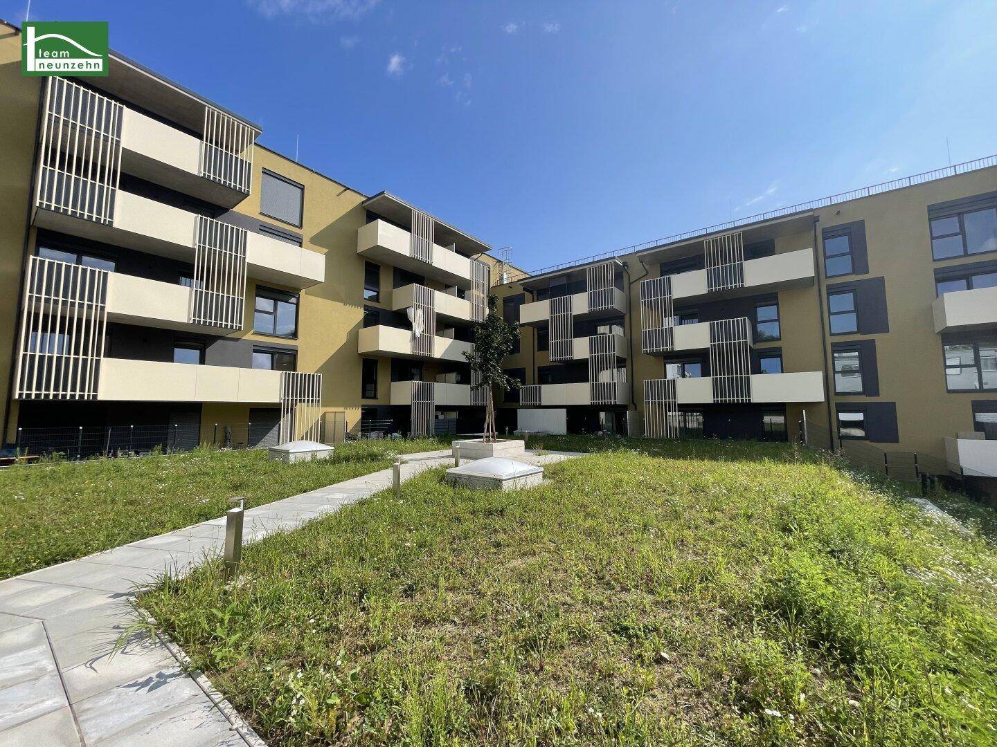 Moderne Wohnanlage mit Balkonen und begrüntem Innenhof unter blauem Himmel.