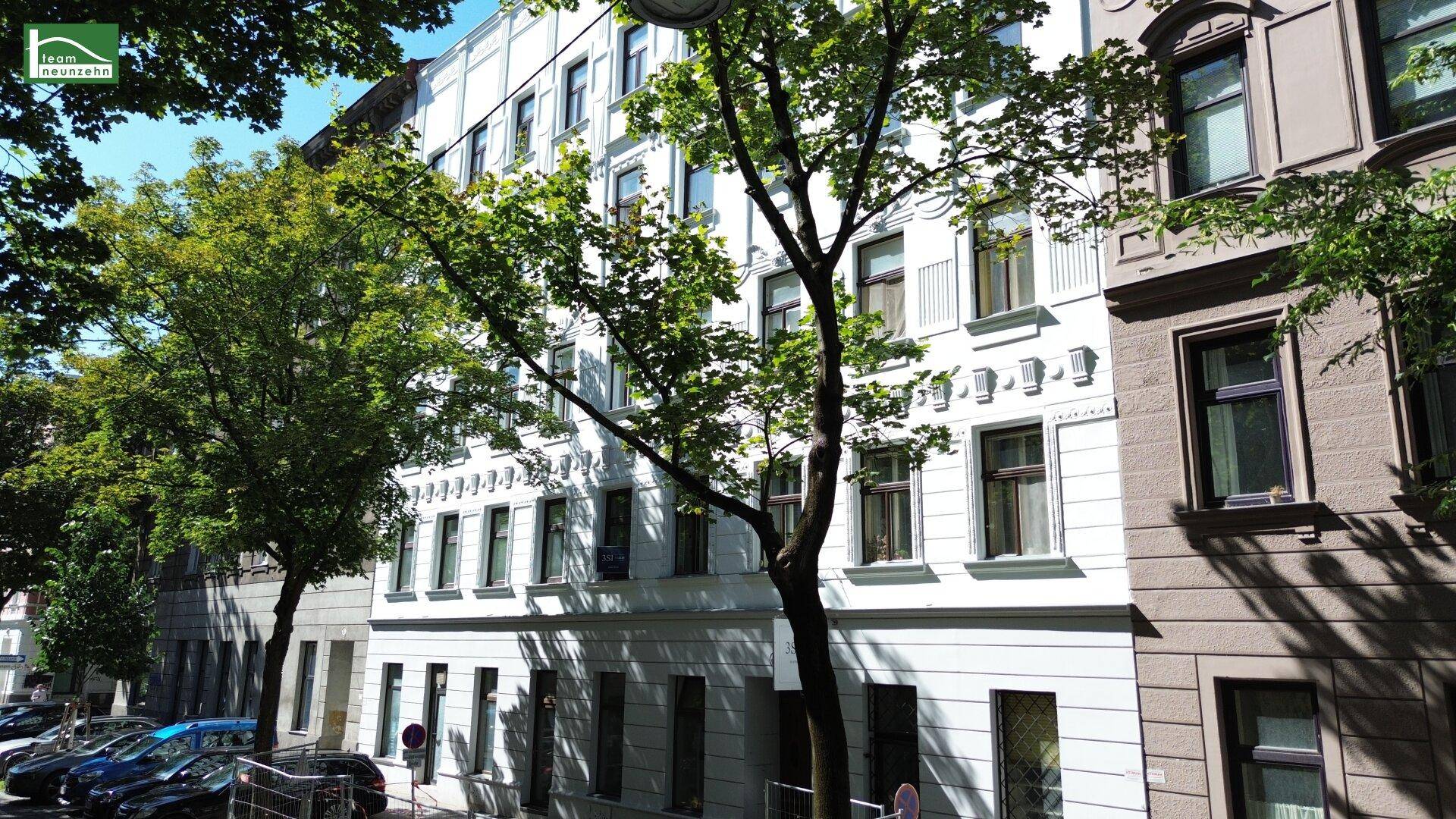 Straßenansicht eines historischen Wohngebäudes mit weißer Fassade, umgeben von Bäumen und parkenden Autos.