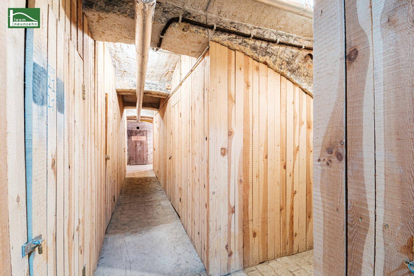 Langer Kellergang mit Holzverschlägen und sichtbaren Rohren an der Decke.