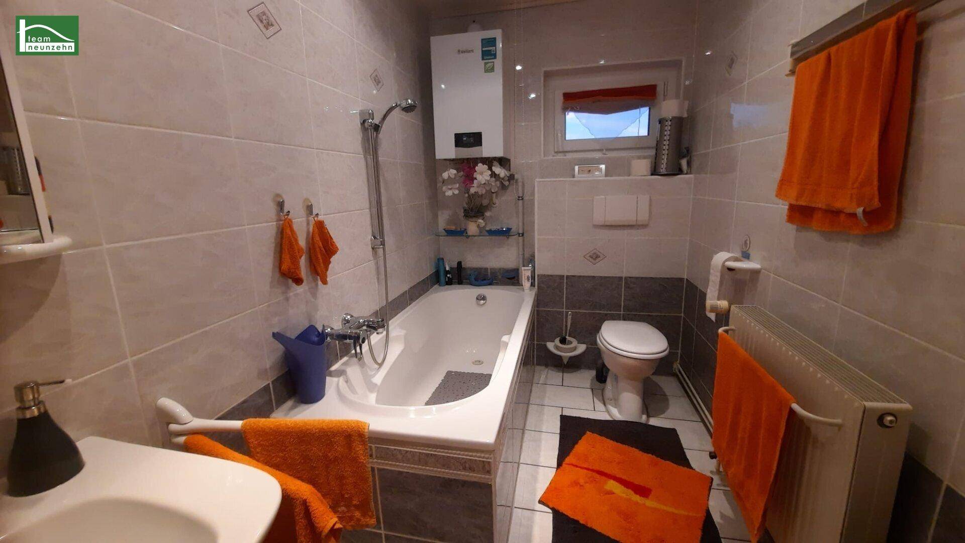 Gefliestes Badezimmer mit Badewanne, Toilette und Fenster, ausgestattet mit orangefarbenen Handtüchern.