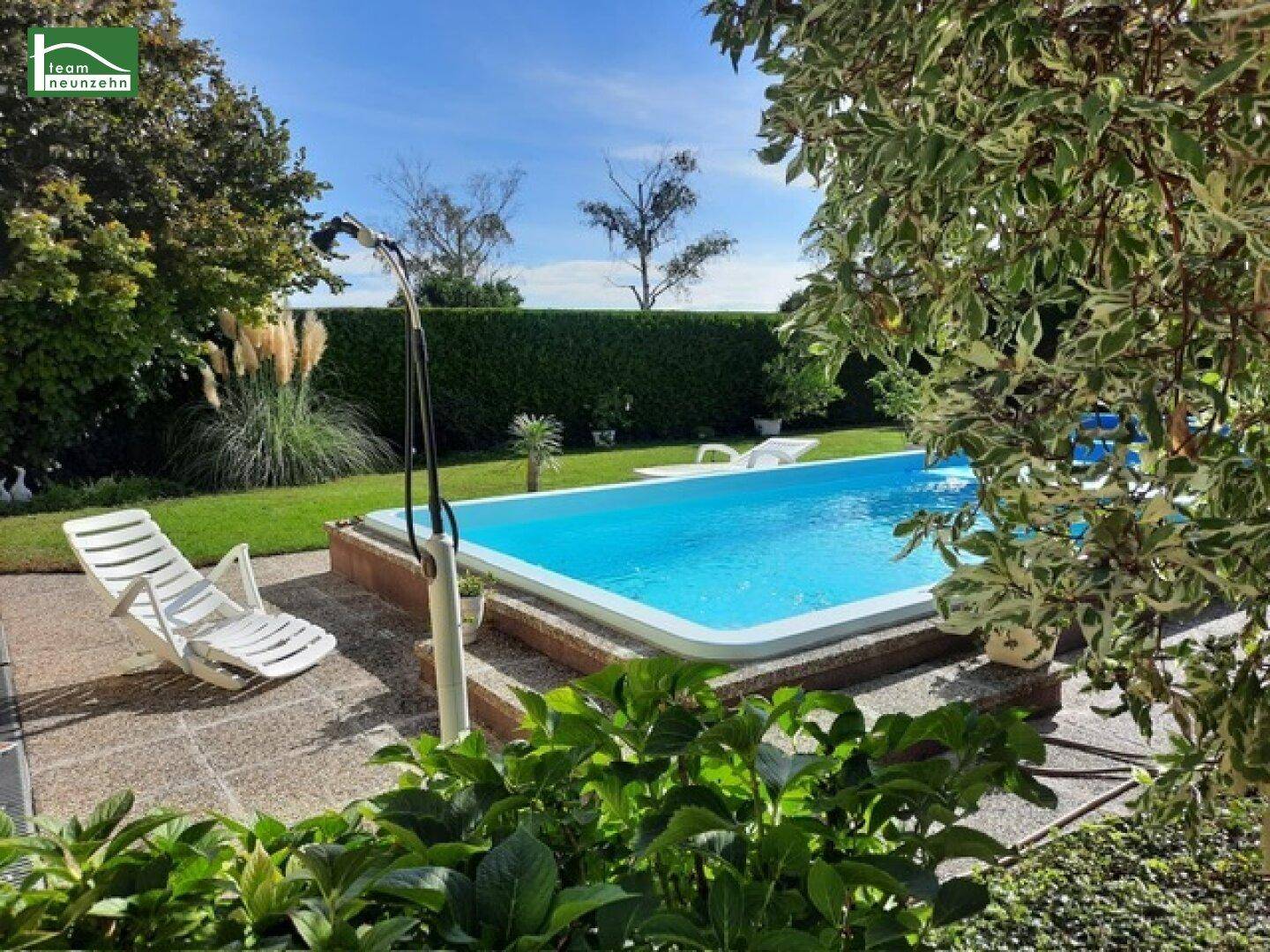 Gepflegter Gartenbereich mit Swimmingpool, Liegestühlen und grüner Hecke unter blauem Himmel.