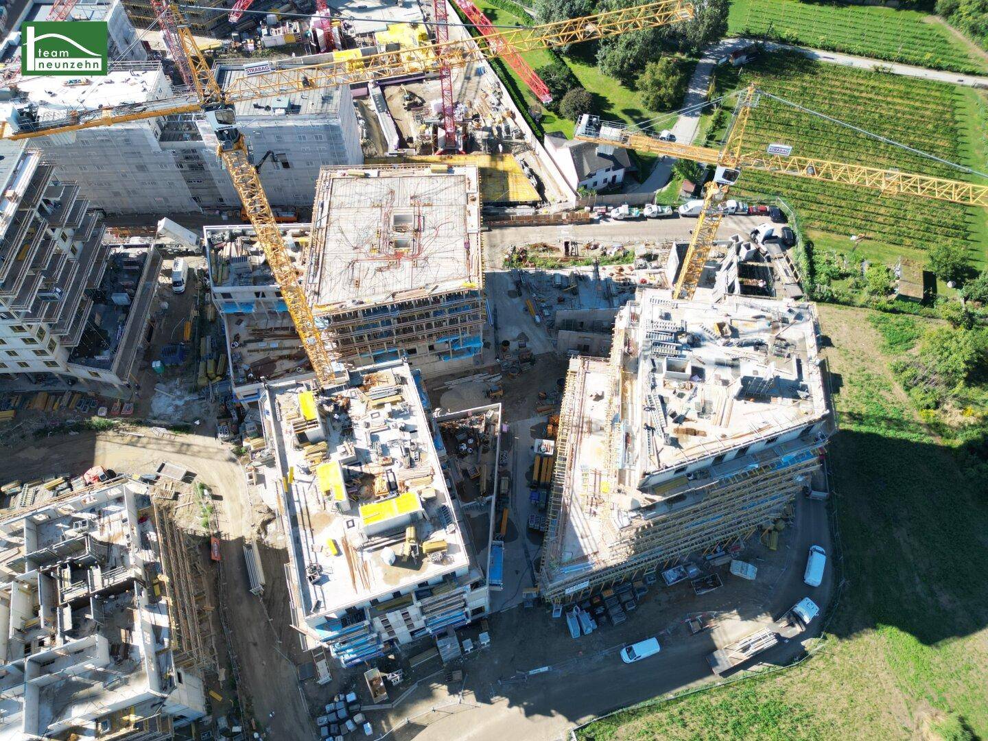 Luftaufnahme einer großen Baustelle mit mehreren im Bau befindlichen Gebäuden und Kränen, umgeben von grünen Feldern und Weinbergen.