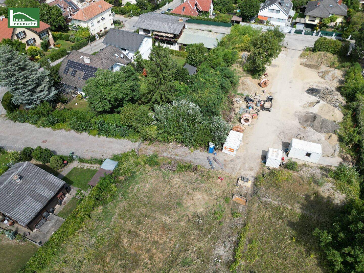 Luftaufnahme eines Baugrundstücks mit umliegenden Wohnhäusern, viel Grün und Baustellenmaterial.
