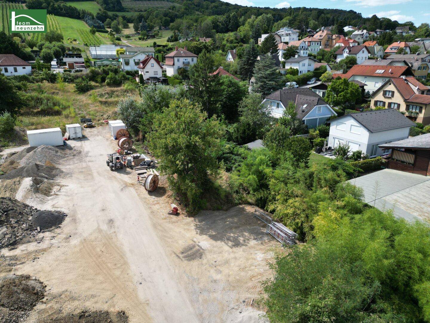 Luftaufnahme einer Baustelle mit umliegenden Häusern und grünen Weinbergen im Hintergrund.
