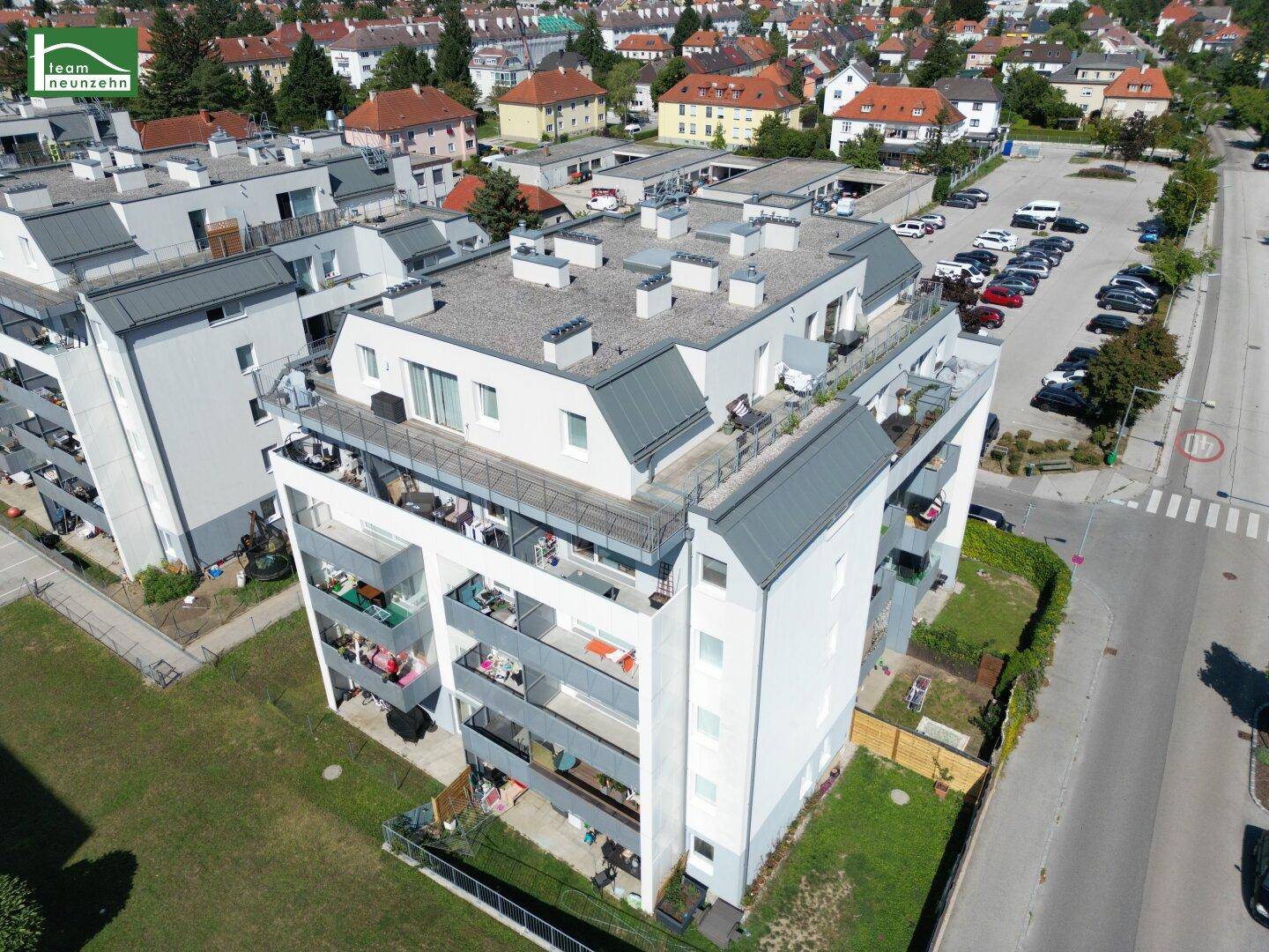 Vogelperspektive auf ein modernes Wohngebäude mit Dachterrassen und Balkonen, umgeben von Straßen und Grünflächen.