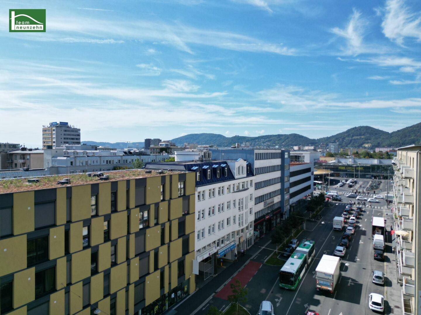 Blick über eine belebte Stadtstraße mit modernen Gebäuden und Bergen im Hintergrund unter blauem Himmel.