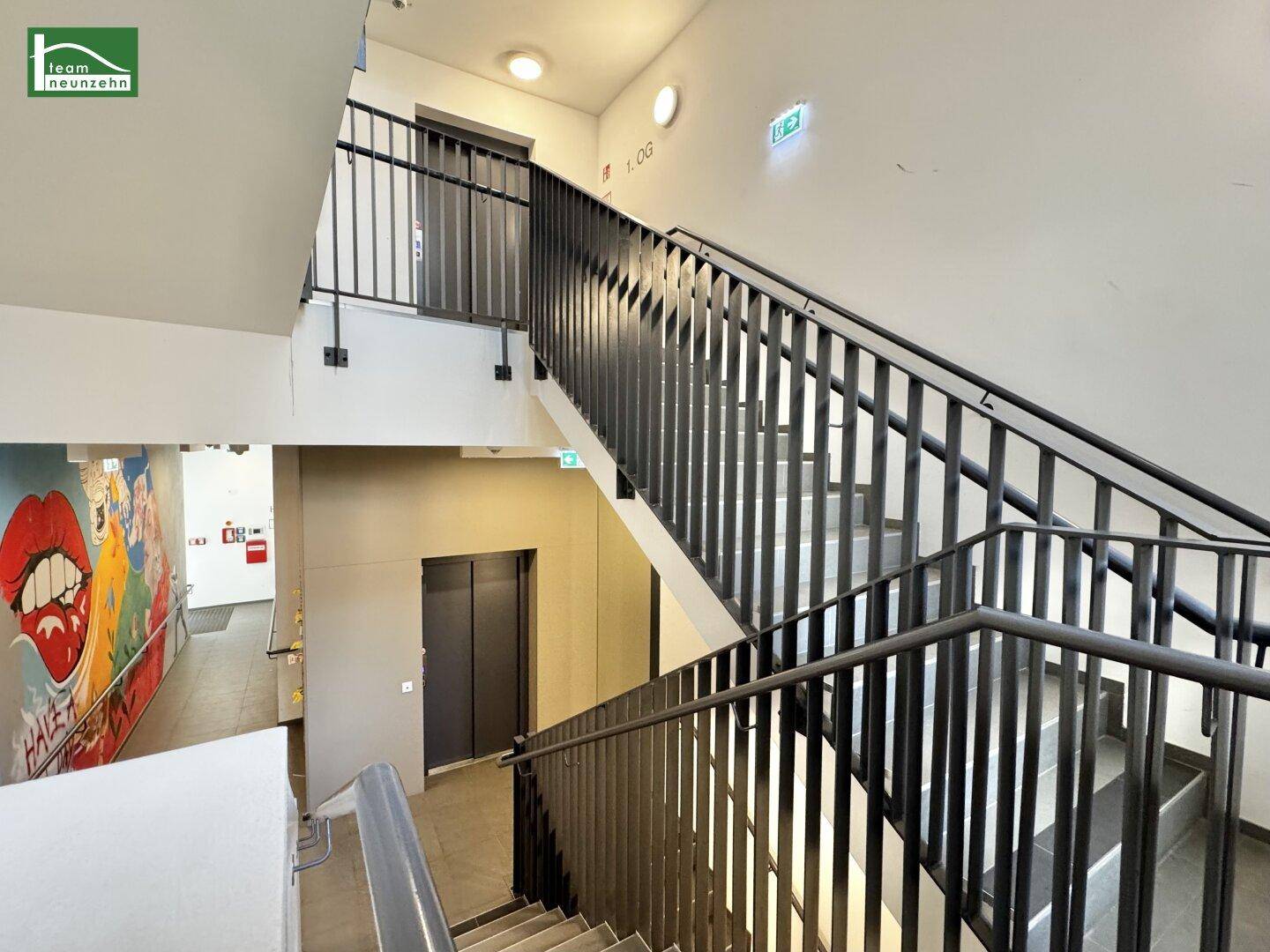 Treppenhaus und Aufzug in einem modernen Gebäude mit künstlerischer Wandgestaltung im Hintergrund.